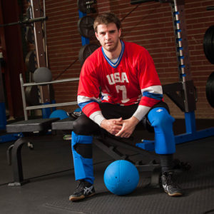 Photo of Matt Simpson dressed in his Team USA Goalball gear and sitting on a bench with a goalball in front of him
